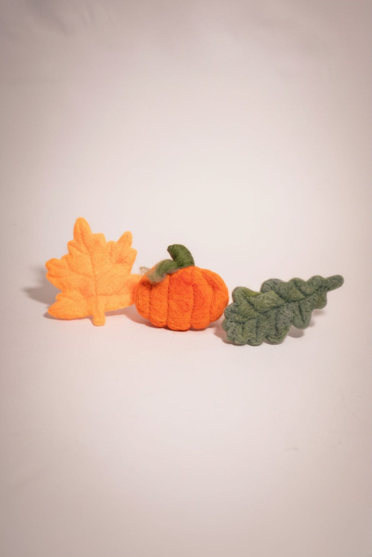 Three felt decorations shaped like a pumpkin, orange leaf, and green leaf on a plain background.