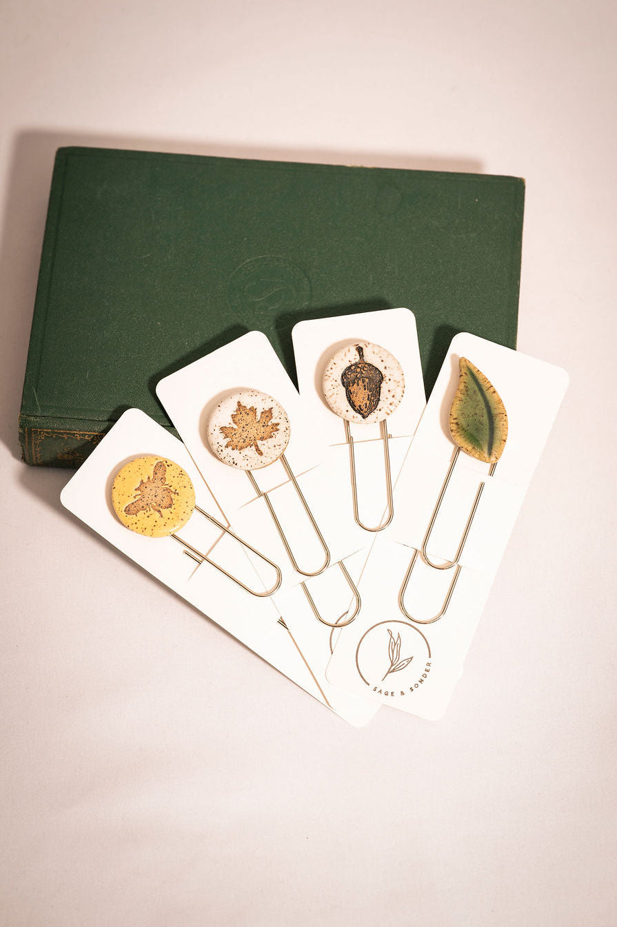 Set of decorative ceramic bookmarks packaged on a white card leaning against a green fabric covered book.