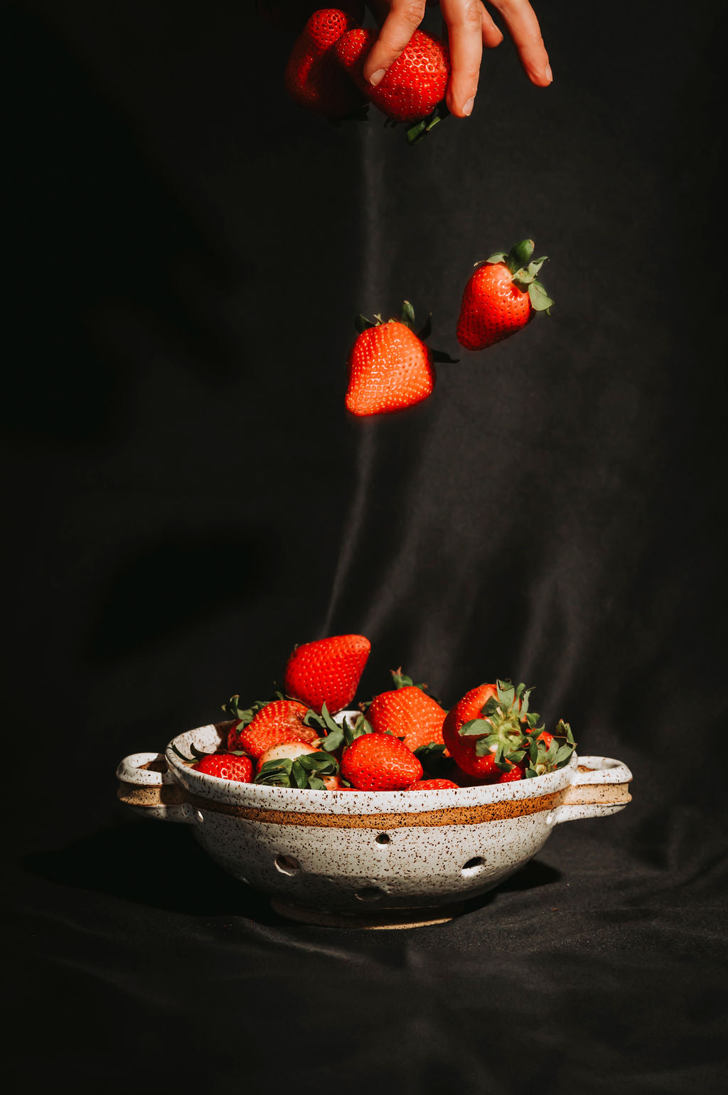 Strawberries falling into a white speckled ceramic colander on a black background
