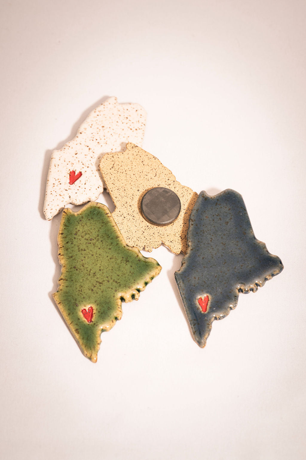 Four ceramic maine magnets shaped in state of Maine with stamped heart designs. One magnet is turned over to see the back.