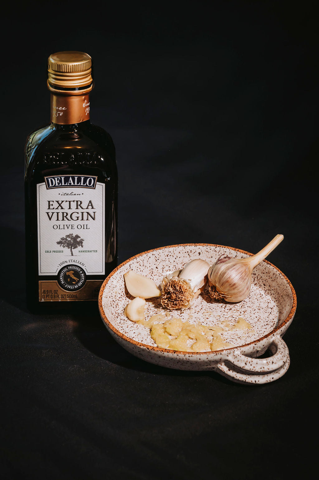 White ceramic garlic grater plate with garlic cloves on the plate and a bottle of olive oil next to it dark background