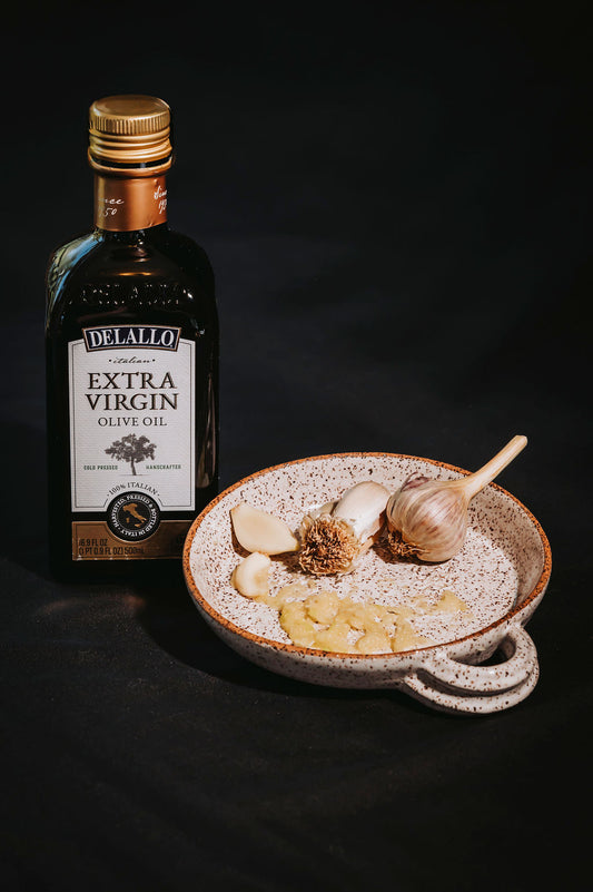 White ceramic garlic grater plate with garlic cloves on the plate and a bottle of olive oil next to it dark background