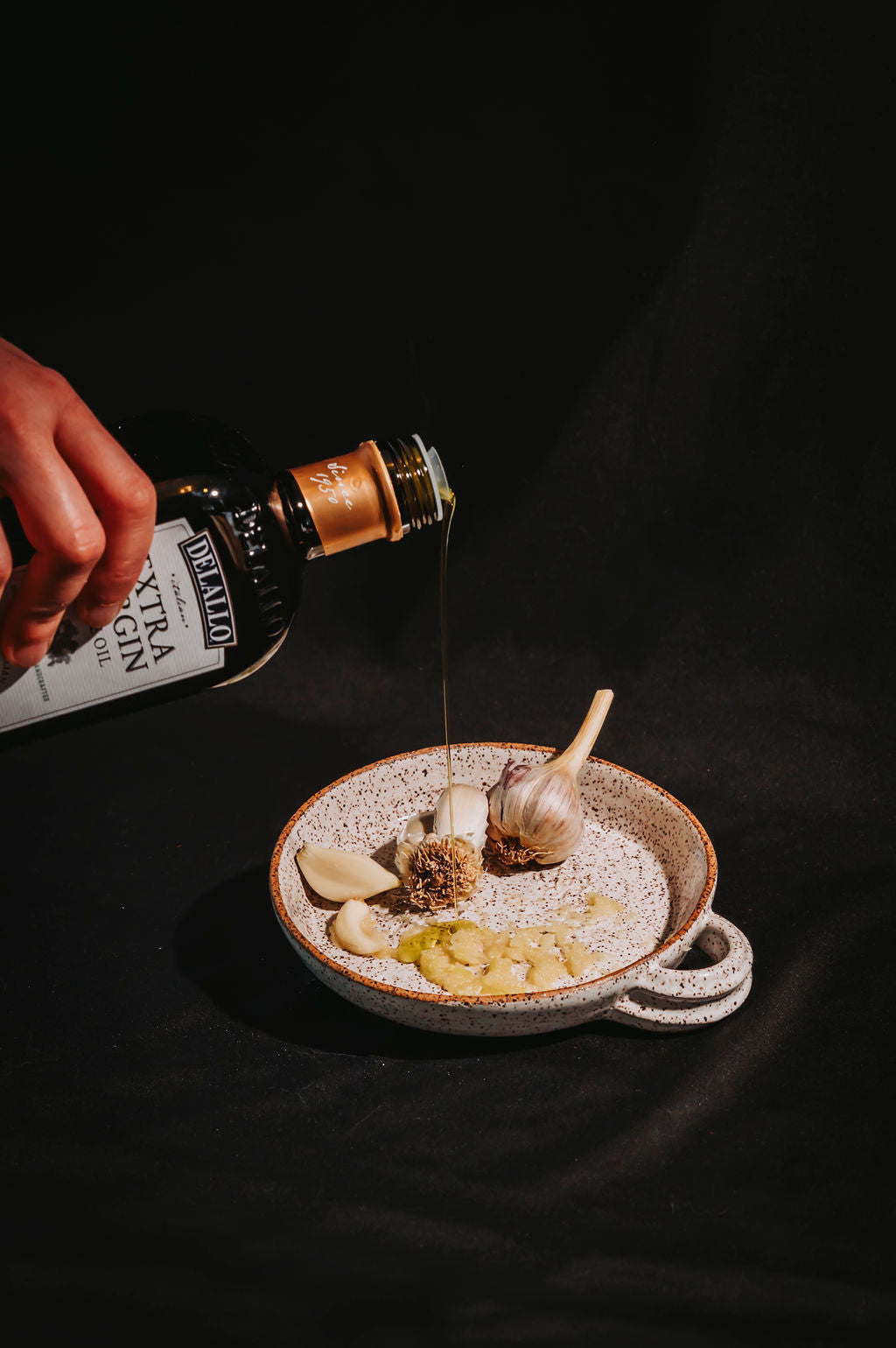 Person pouring olive oil from a bottle onto a ceramic garlic grater that has garlic and herbs on it