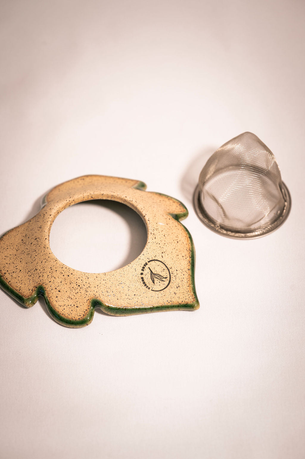 Back of a ceramic tea infuser in the shape of a leaf with a silver mesh tea strainer basket sitting next to it.