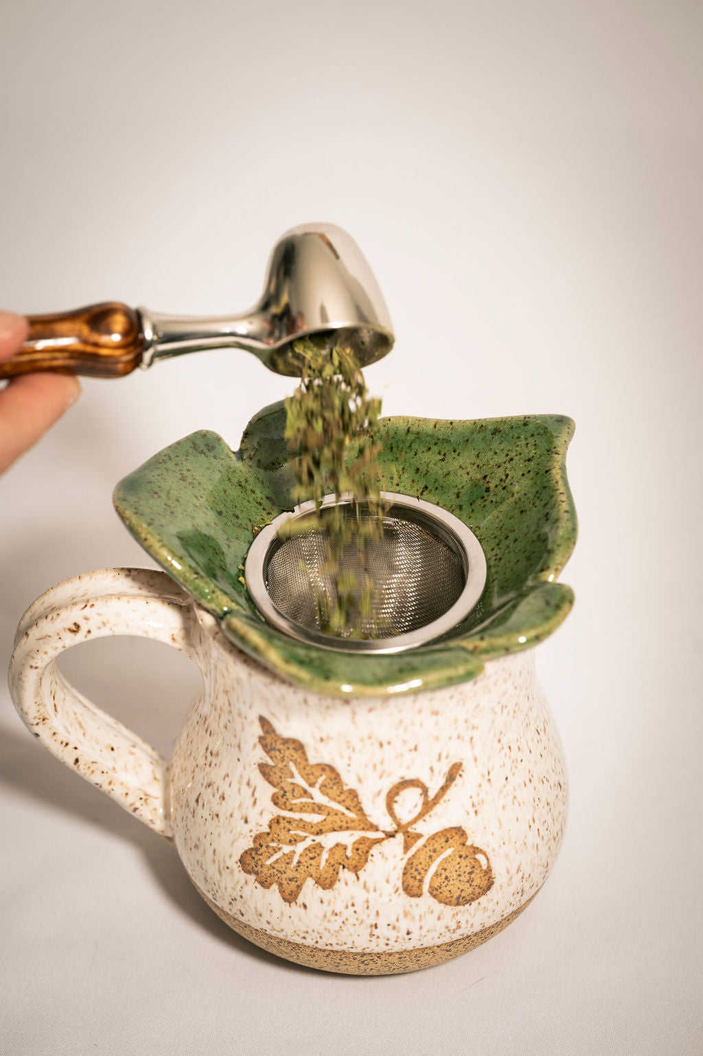 Ceramic tea infuser with a leaf design being used to pour green tea leaves into it.