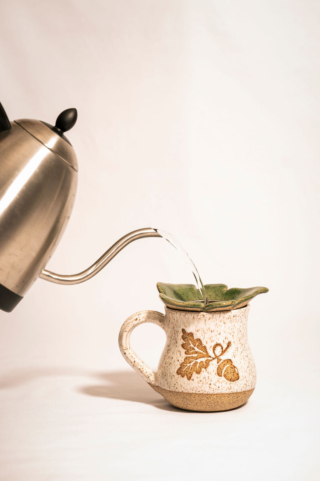 Tea kettle pouring water into a speckled mug with an acorn design with a green ceramic tea strainer resting on top.
