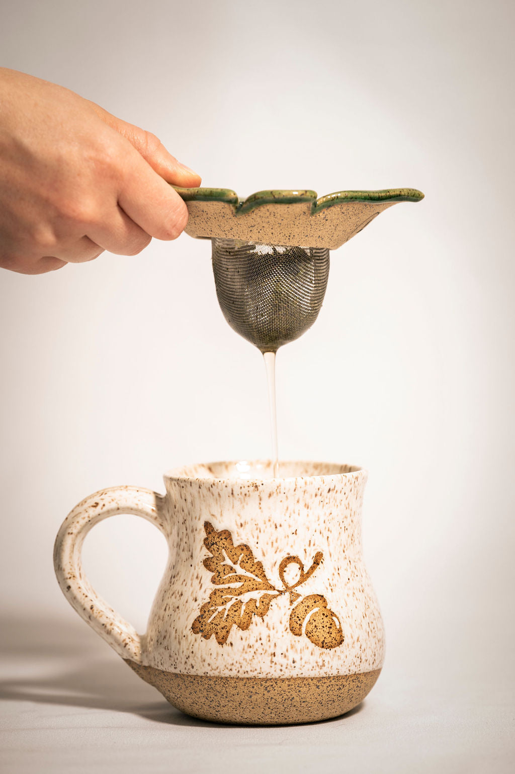 Person lifting a ceramic tea strainer from a speckled mug with an acorn design.