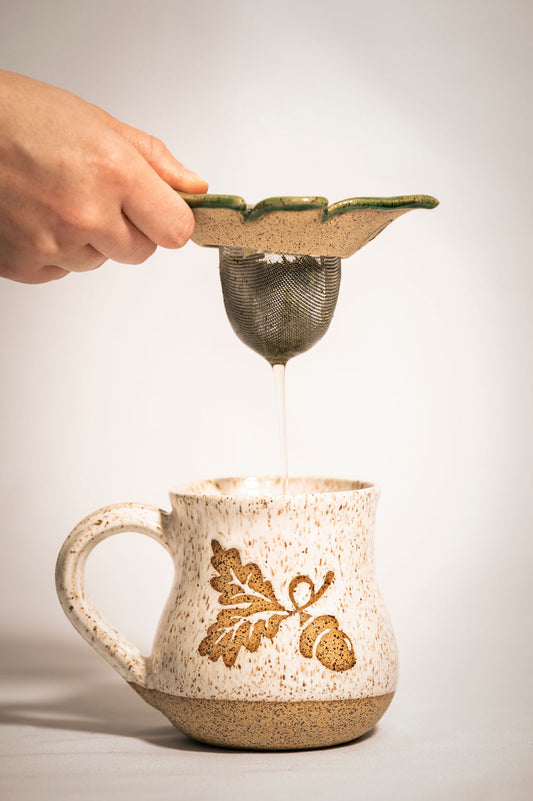 Person lifting a ceramic tea strainer from a speckled mug with an acorn design.
