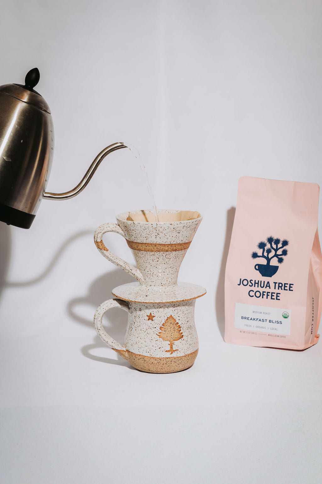 Ceramic coffee pourover sitting on a mug with a water kettle and 'Joshua Tree Coffee' bag nearby on a white background