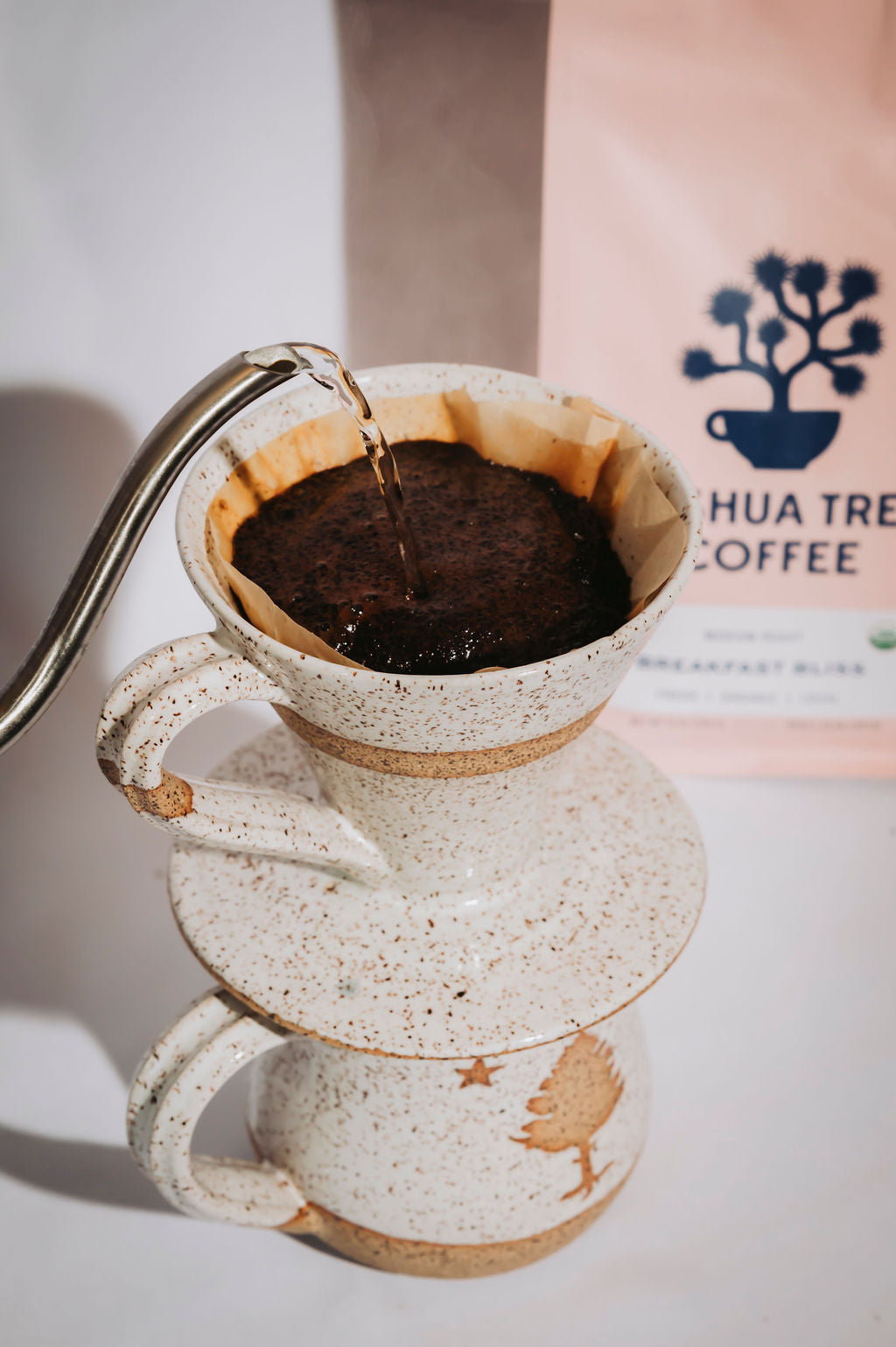 Ceramic pour-over coffee maker with hot water being poured into it, with a coffee bag in the background.