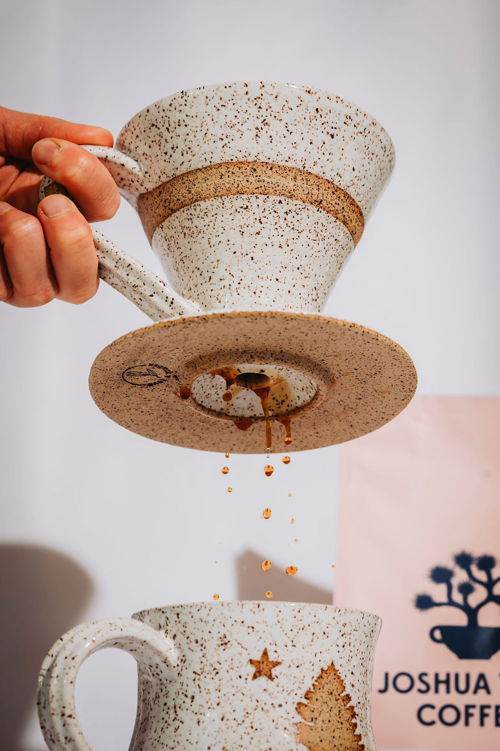 Person picking up a coffee pour over from a speckled ceramic mug. Shows coffee brewing and dripping into the matching mug.