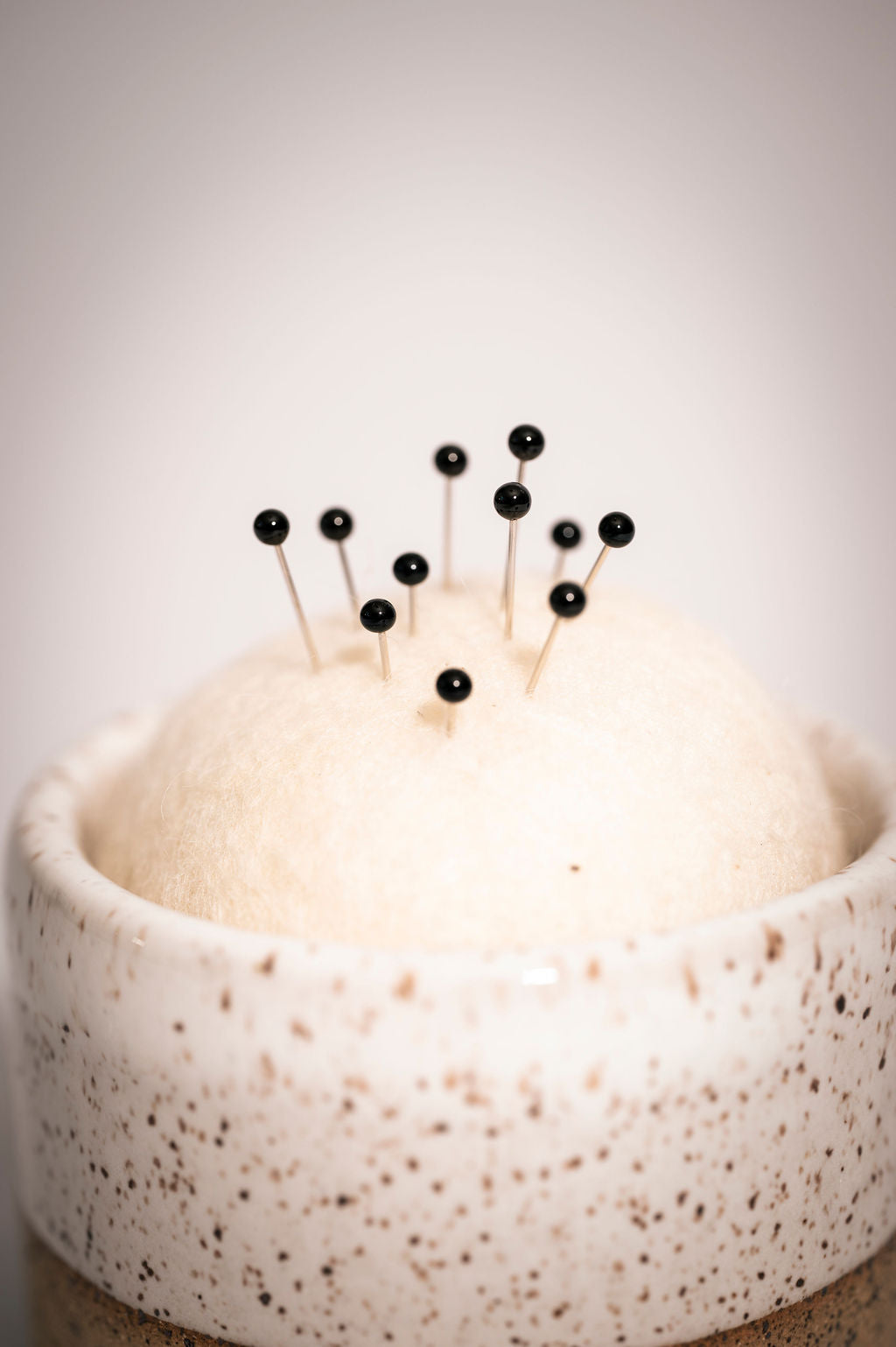 Close up of White speckled ceramic tumbler with a wool pin cushion and black pins sticking out.