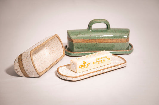 Two ceramic butter dishes. The front speckled white butter dish has a lid laying beside it and shows a stick of butter on the tray.
