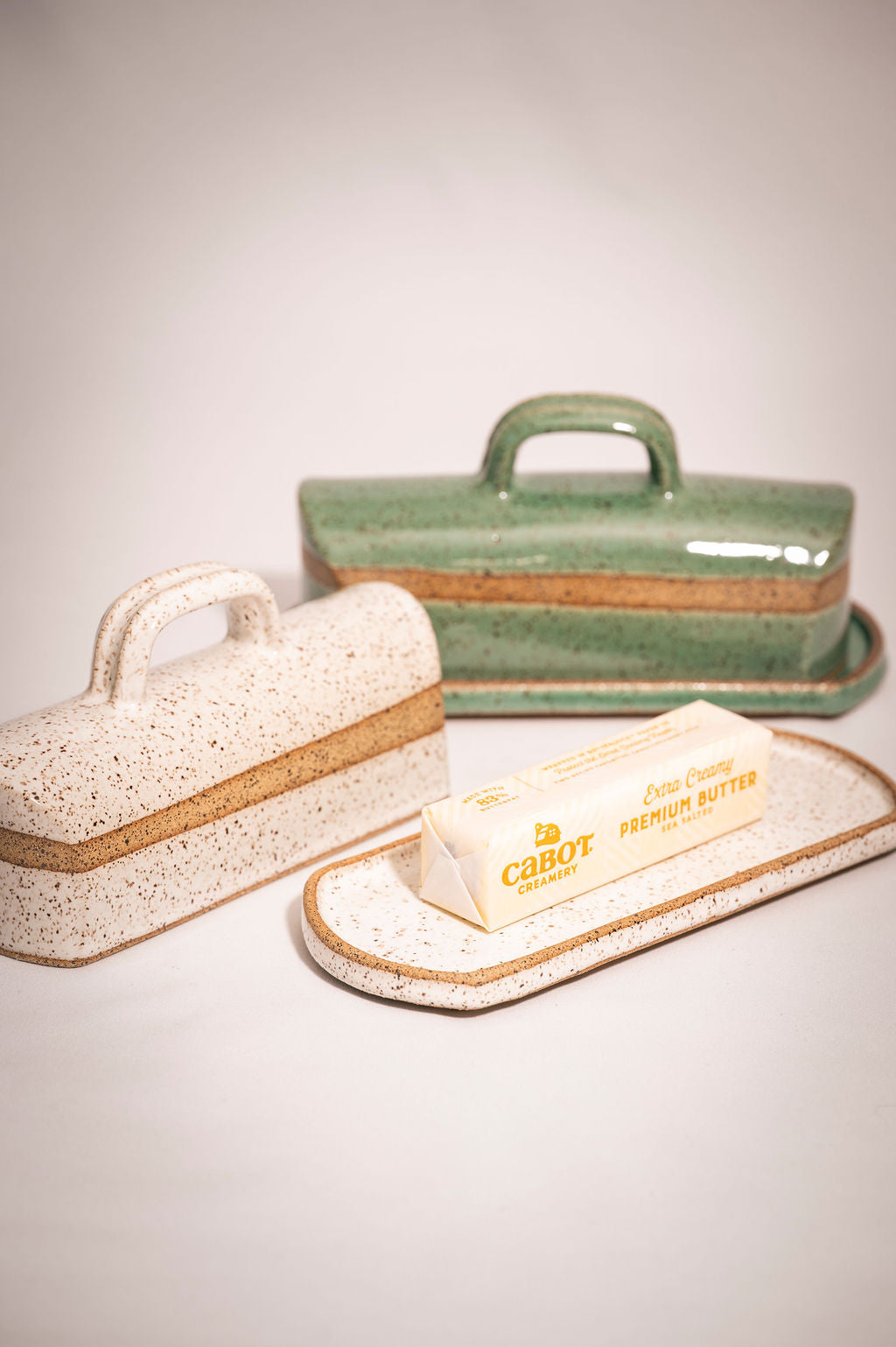 Two ceramic butter dishes. The front speckled white butter dish has a lid sitting beside it and shows a stick of butter on the tray.