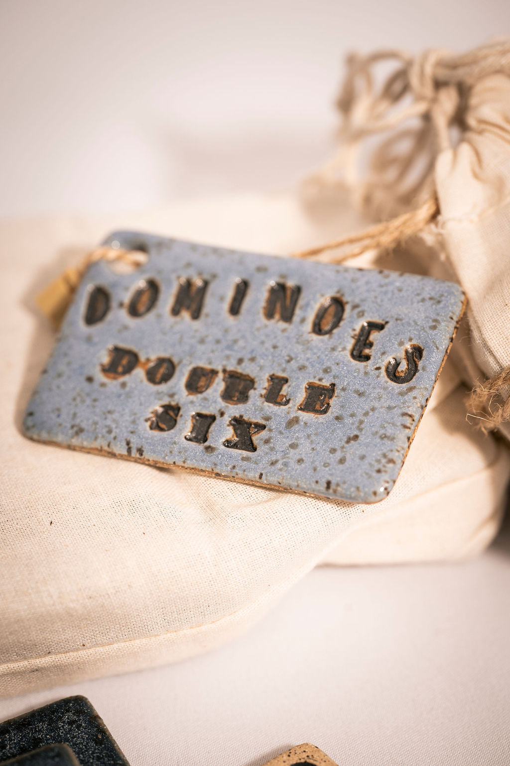 Light blue rectangular ceramic tag with text reading "Dominoes Double Six" on a beige fabric background