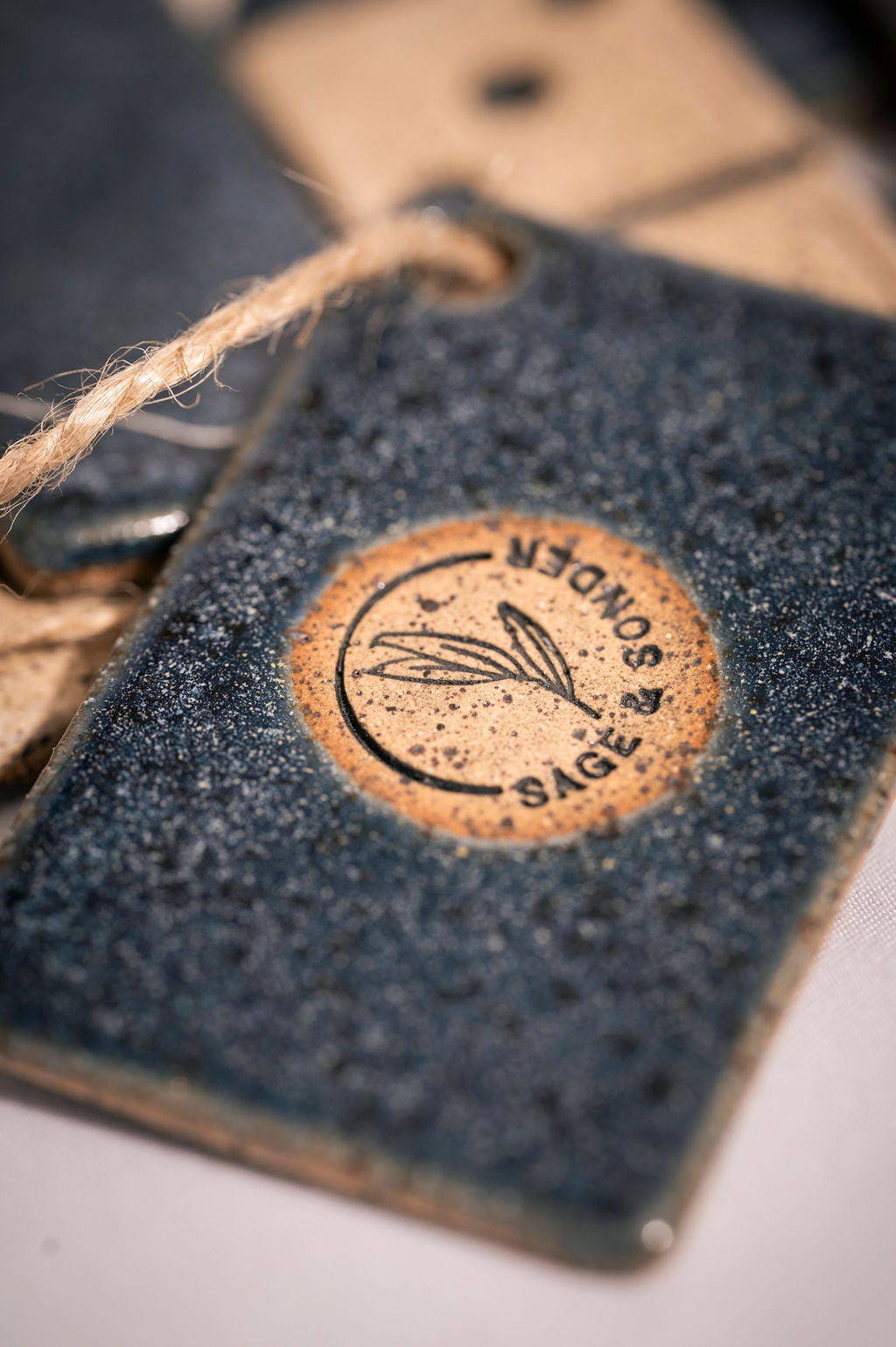 Clsoe up of dark denim blue tag with a brown circular logo and string, featuring the brand 'Sage & Sonder'